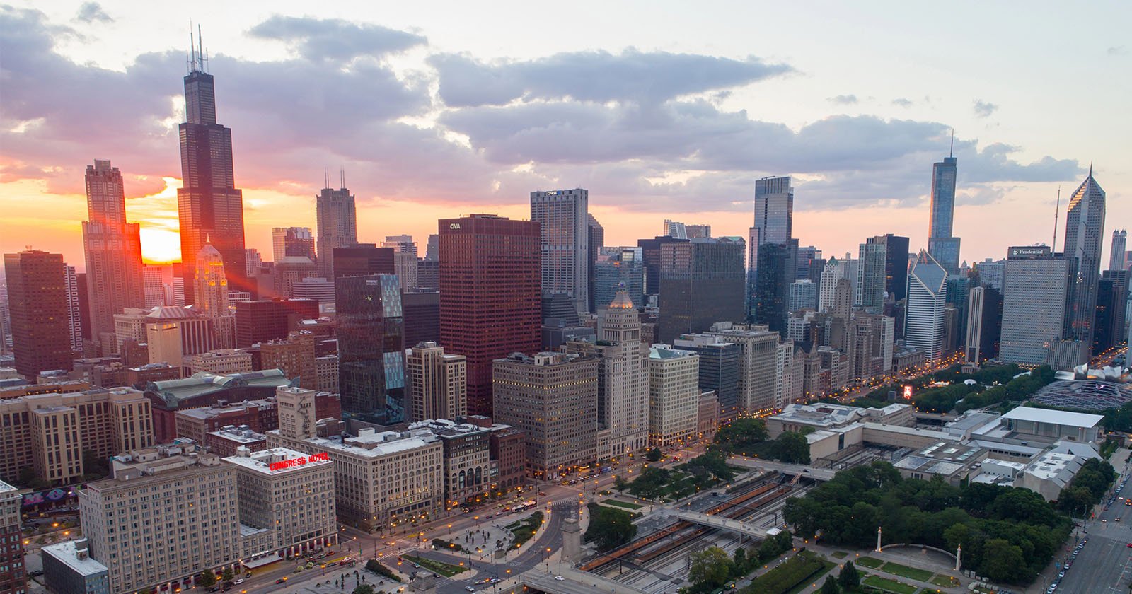 Aerial view of a city skyline at sunset, with sunlight reflecting off tall skyscrapers and clouds in the sky. Streets and green spaces are visible in the foreground.