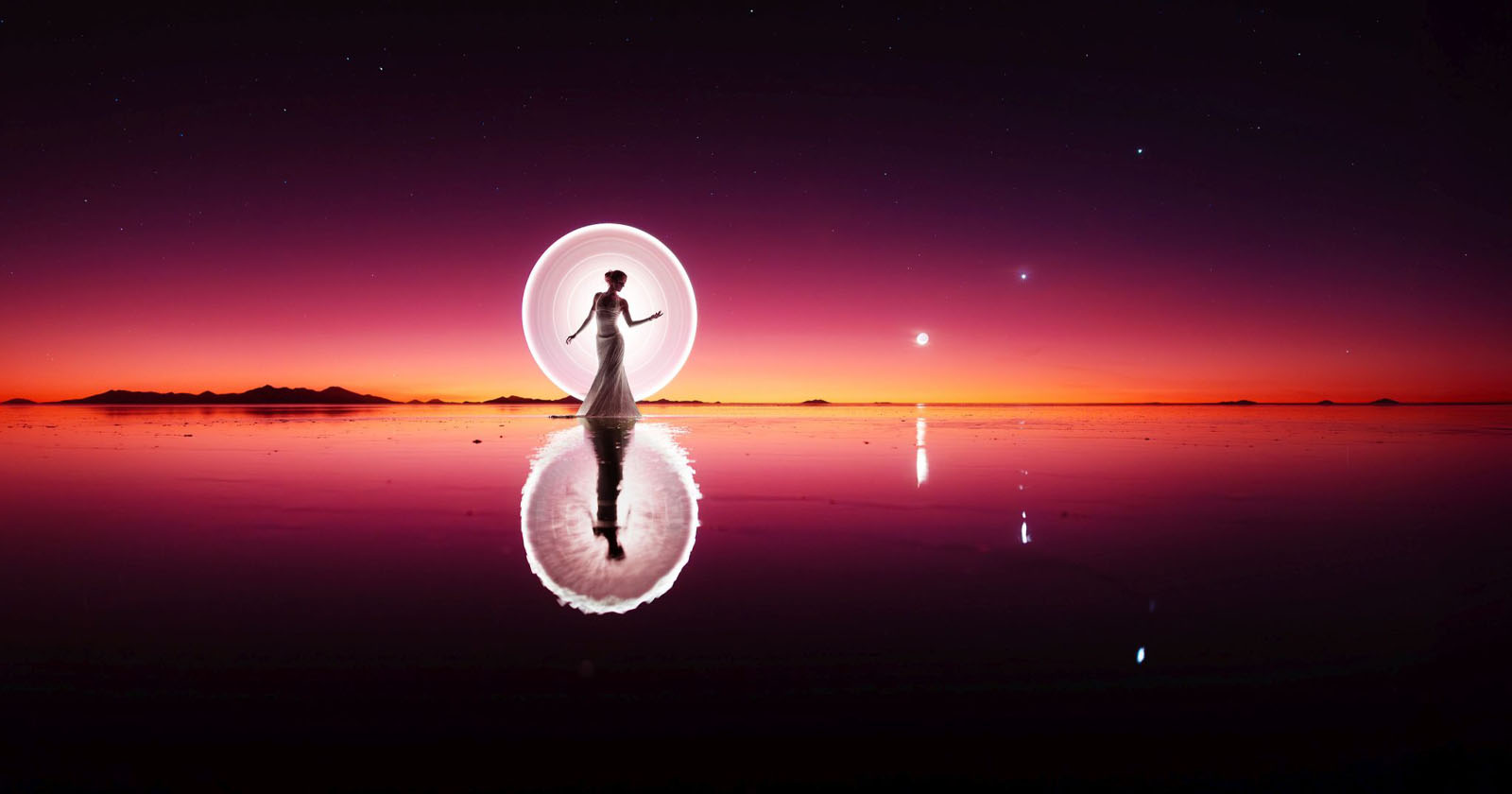 A silhouetted person in a flowing dress stands on a reflective, water-covered surface. Behind them, a glowing circle of light creates an ethereal effect. The sky is a gradient of deep purple to orange as the sun sets, with a few stars visible above.