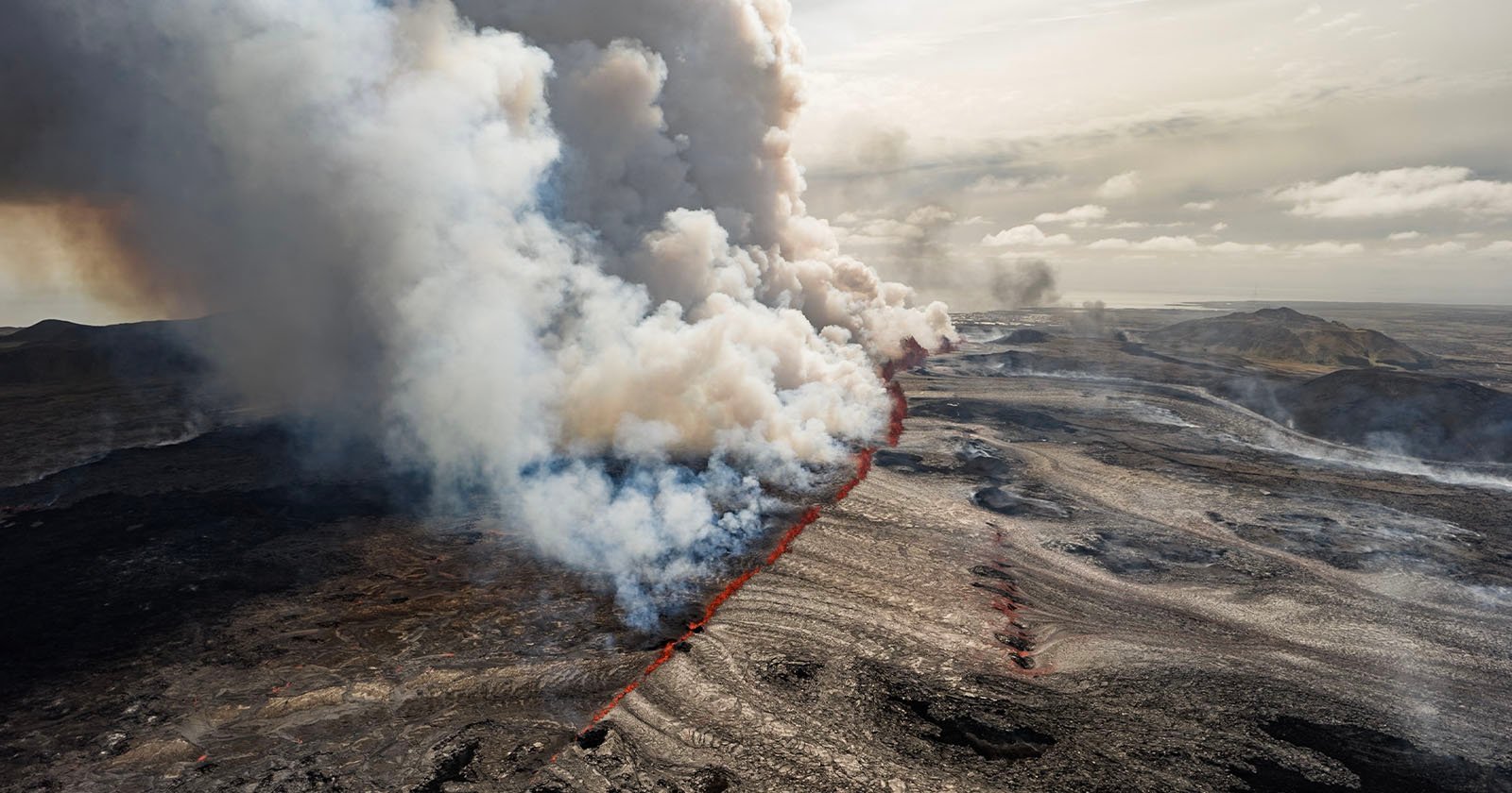 Aerial view of an active volcanic fissure emitting large plumes of white and gray smoke, with a bright red flow of lava extending through a rugged, rocky landscape. The sky is overcast, casting a muted light over the scene.