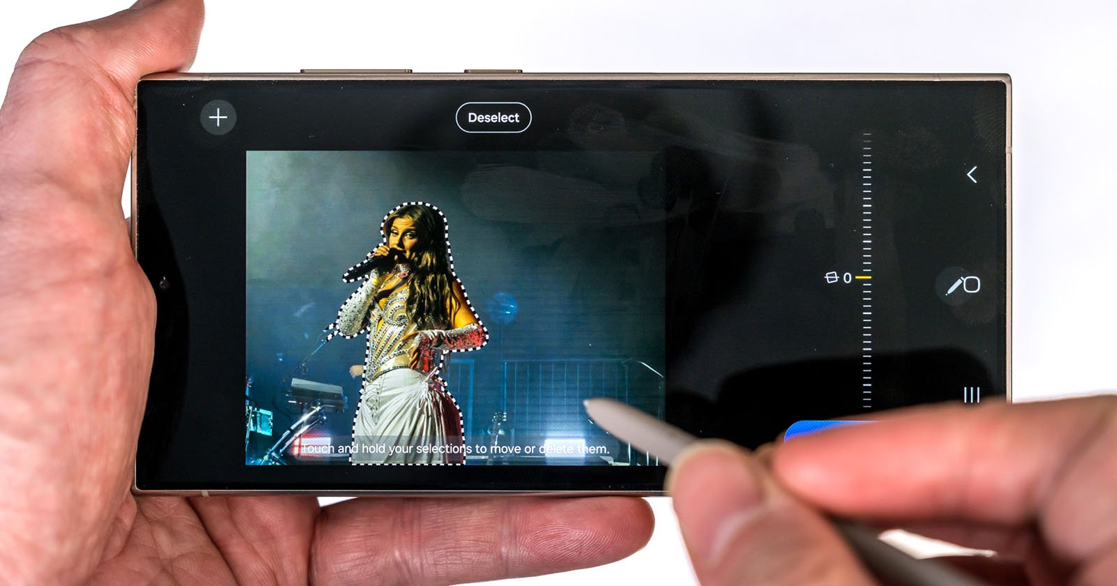 A hand holding a smartphone with a stylus, editing an image of a female singer on stage, highlighting her figure with a selection tool on the screen.