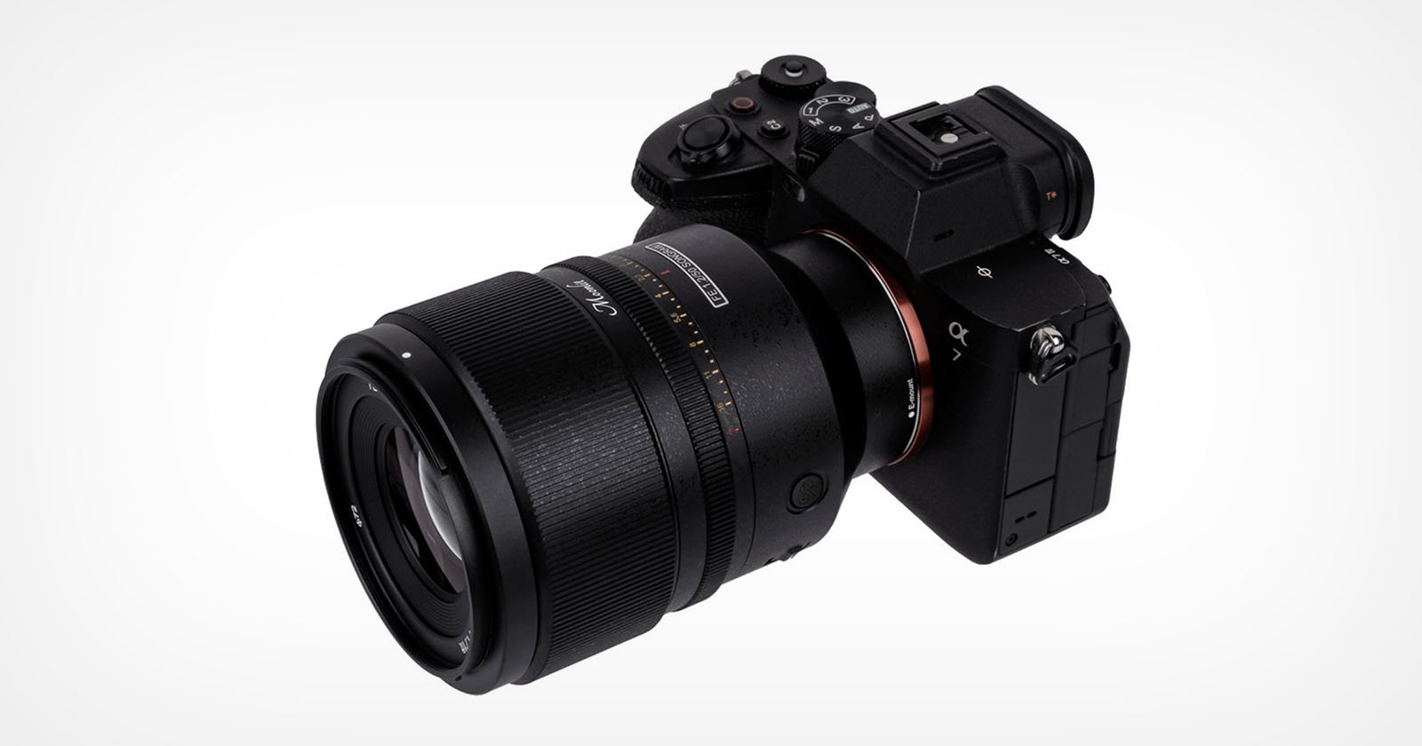 A black digital camera with a protruding lens is positioned at an angle against a plain white background. The camera features several buttons and a textured grip, with some visible markings and logos on the body and lens.