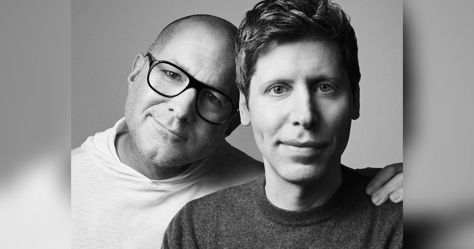 Two men pose closely together for a black and white portrait. The man on the left is bald, wears glasses, and leans his head on the other man's shoulder. The man on the right has short hair and wears a sweater, looking directly at the camera.