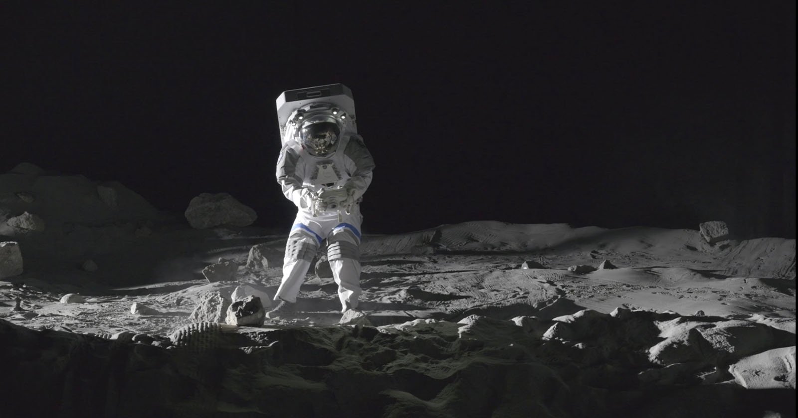 An astronaut in a spacesuit stands on a rocky, dusty lunar surface with scattered rocks and shadows, against the blackness of space.