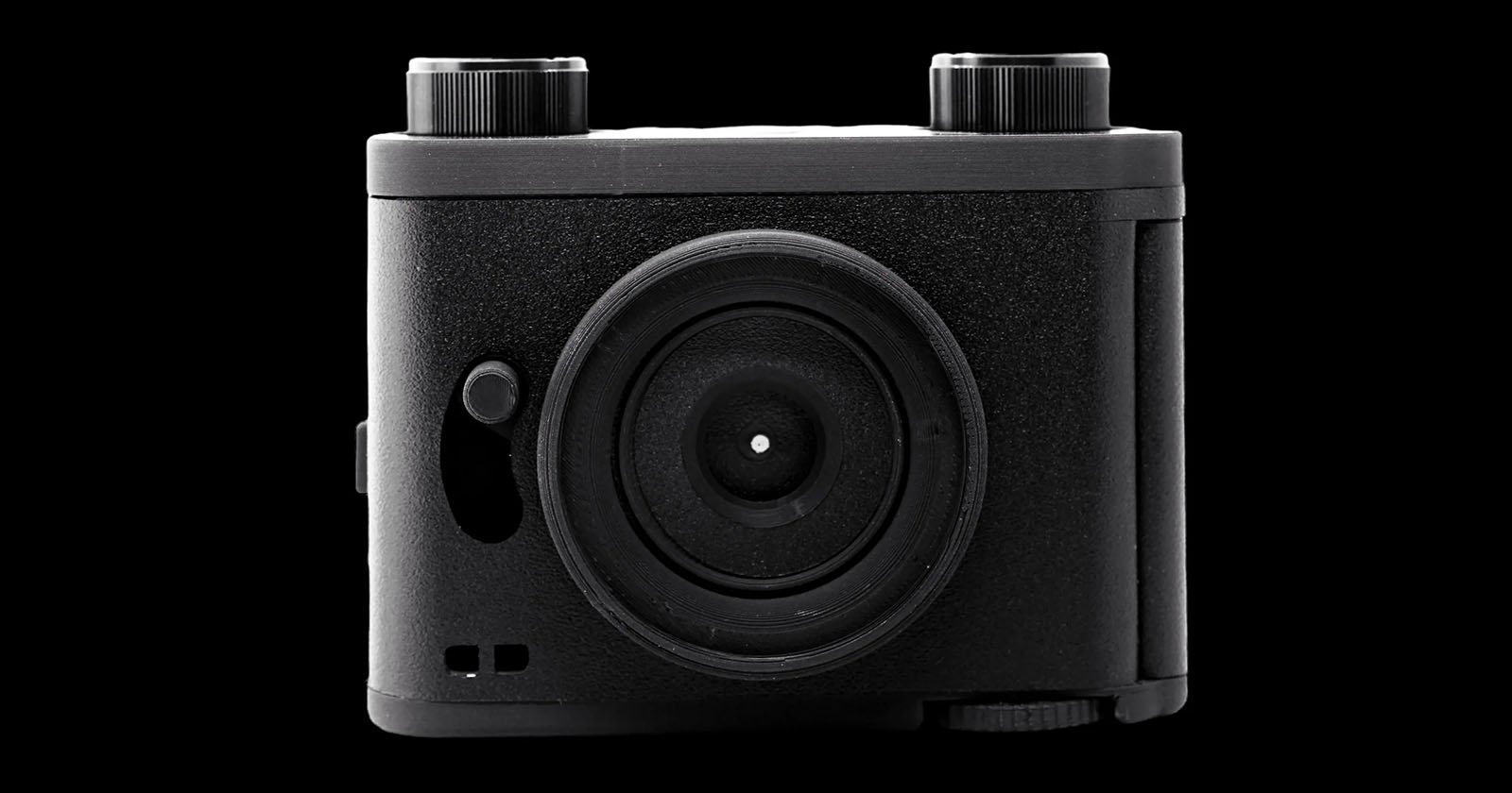 A black, rectangular film camera with a textured finish is centered against a solid black background. It features a large circular lens in the middle, two dials on the top, and a small switch on the front left side. The overall design is minimalist and compact.