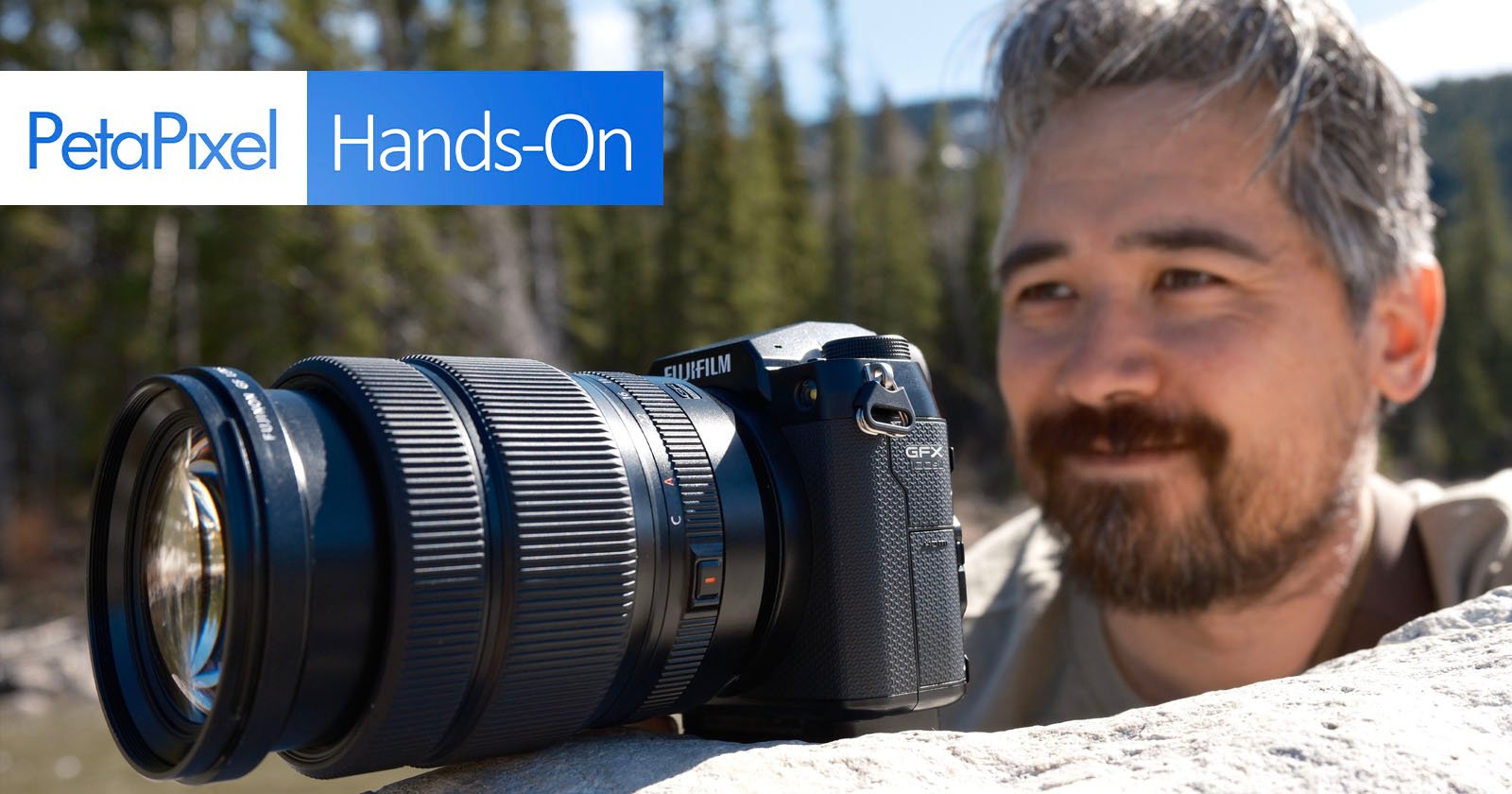A close-up shot of a Fujifilm GFX100S camera with a large lens resting on a rock, with a blurred background of trees and water. The PetaPixel logo and the words Hands-On are displayed in the top left corner. A person with a beard is looking at the camera.