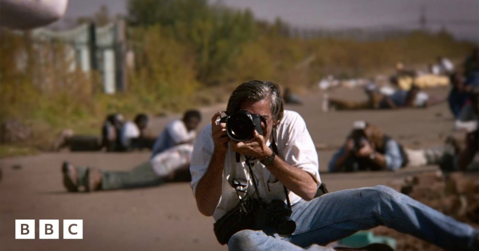 A photographer sits on the ground aiming his camera at the viewer, while several people and photographers are lying on a road in the background. The BBC logo appears in the lower left corner.