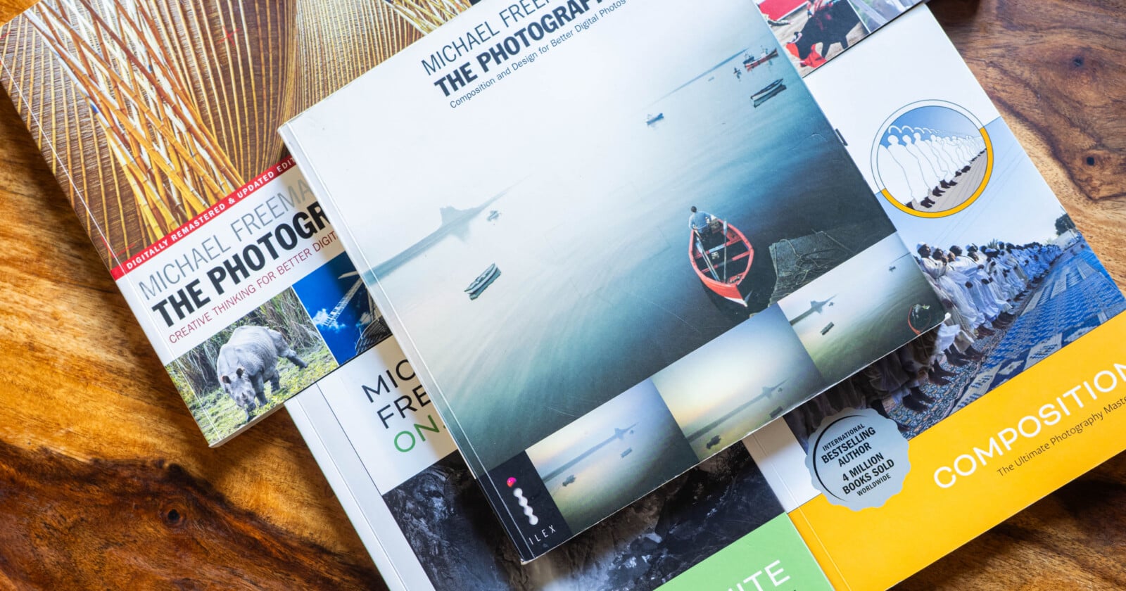 A collection of photography books by Michael Freeman are spread out on a wooden surface, with colorful covers featuring images of landscapes, animals, and people. The central book displays a serene water scene with a red boat.