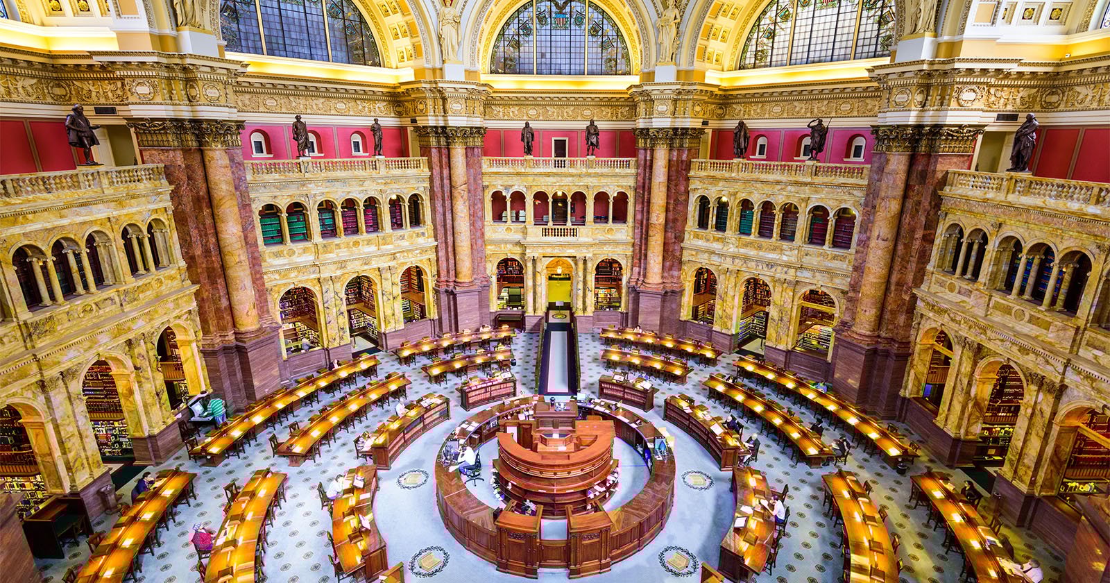 A grand, ornate reading room with arched windows and tall bookshelves, featuring a circular wooden desk at the center and rows of desks with lamps arranged in a semi-circle beneath a domed ceiling.