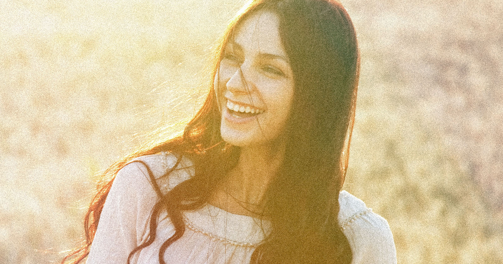 A woman with long dark hair wearing a white blouse smiles brightly in soft, golden sunlight, with an out-of-focus natural background.
