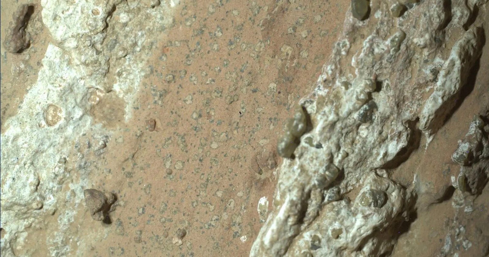 Close-up view of a rough, sandy surface with light brown and tan tones, scattered small stones, and irregular patches of white, textured material running diagonally across the image.