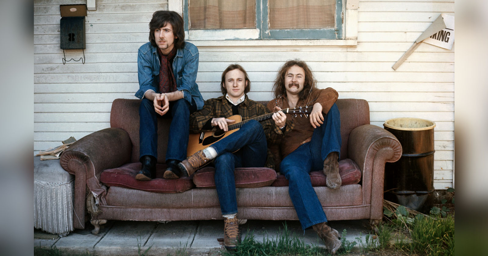 Three men sit on a worn-out couch outside a house. The man on the left has long hair and is wearing a denim jacket, while the middle man holds an acoustic guitar. The man on the right has long hair and a mustache. There's an old barrel and a sign that reads PARKING nearby.