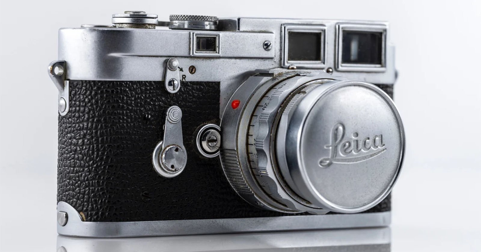 A close-up of a vintage Leica camera with a silver body and black textured grip. The lens cap prominently displays the Leica logo. The camera rests against a white background.