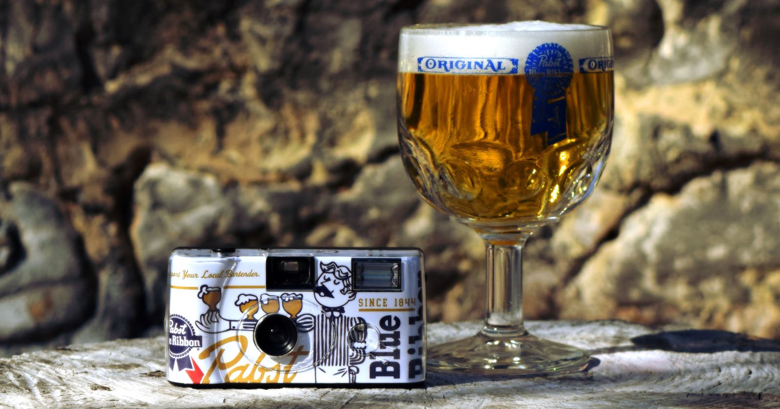 A vintage camera with a Pabst Blue Ribbon design is placed on a surface beside a glass of golden beer with a foamy head. The beer glass is labeled Original. The background features a stone wall with sunlight casting warm tones on the scene.