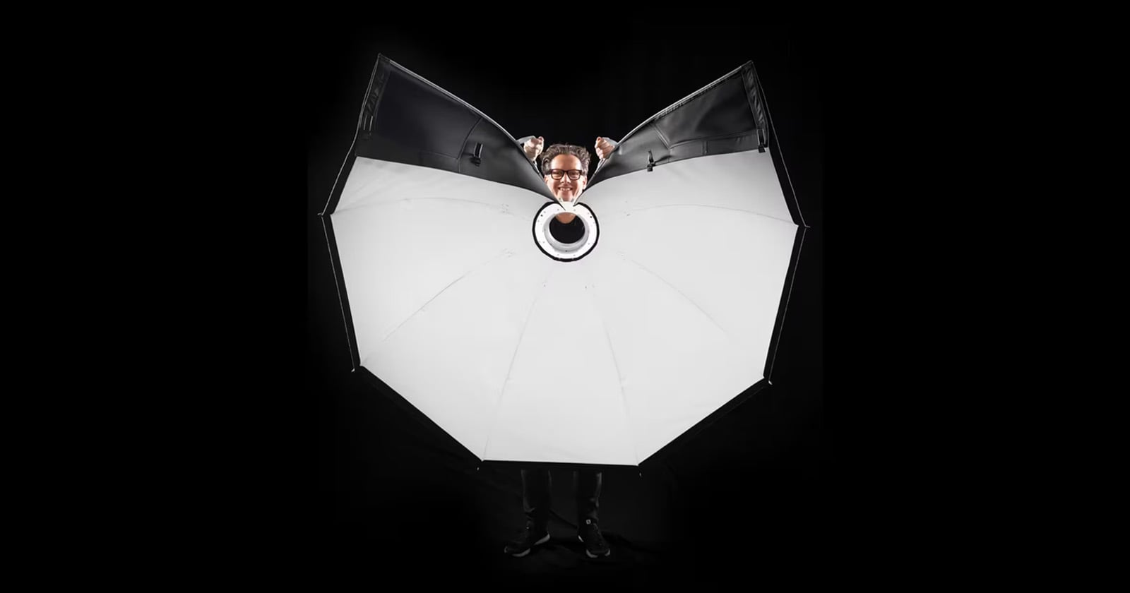 A person stands behind a large, open, white photography umbrella with only their head and hands visible above it, against a black background.