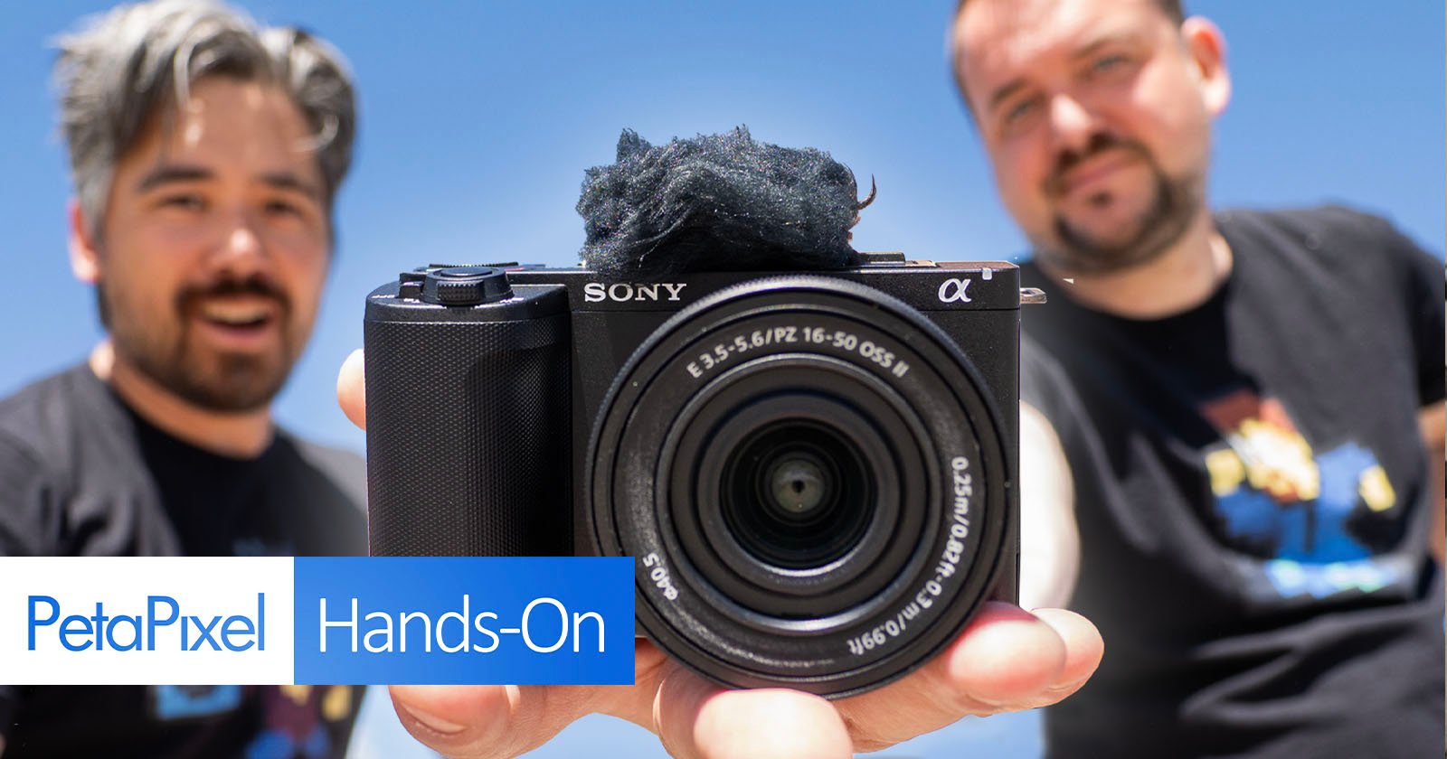 Two individuals are holding a Sony camera up to the lens with a backdrop of a clear blue sky. The image includes the text PetaPixel Hands-On in the bottom-left corner. The camera has a lens with visible specifications, and one person slightly blurred in the background.