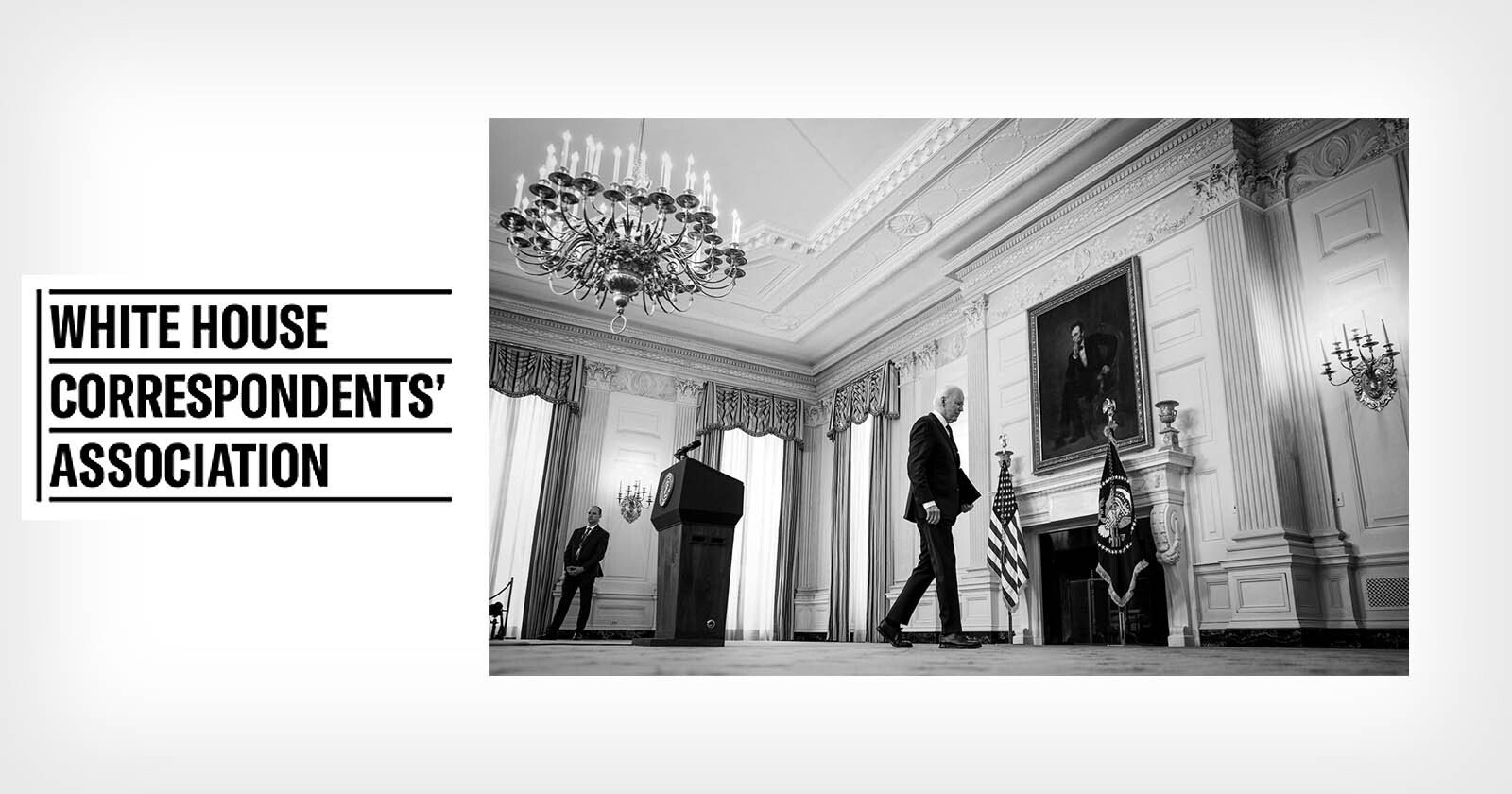 Black and white photo of a man walking past a fireplace and portrait in an ornate room with chandeliers; podium on left. Text on left reads White House Correspondents' Association.