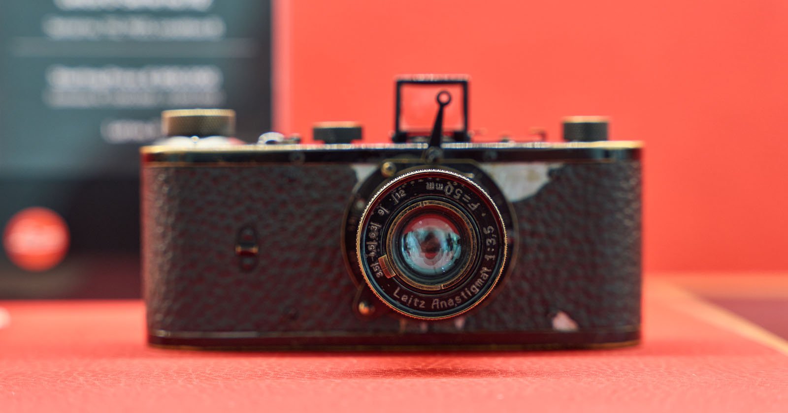 A vintage Leica camera with a textured black body and a round lens is displayed against a red background. The lens reads Leitz Anastigmat. The camera shows visible signs of wear.