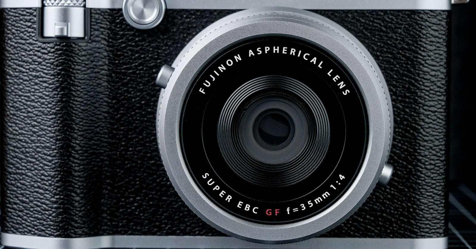 Close-up of a vintage-style camera lens with a black and silver body. The lens is labeled Fujinon Aspherical Lens and Super EBC GF f=35mm 1:4. The camera has a classic textured grip surrounding the lens.