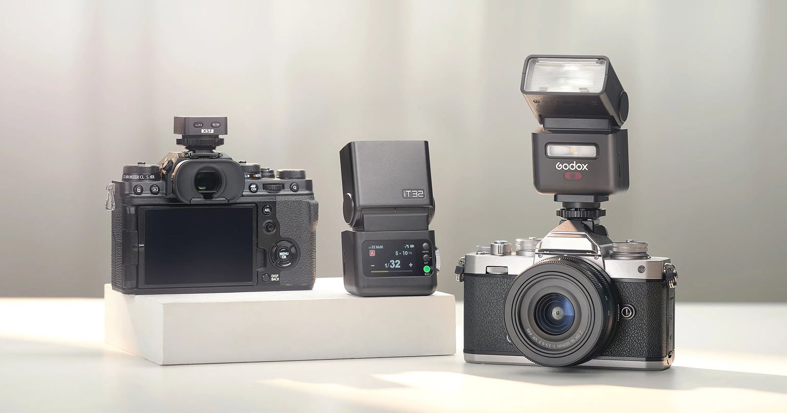 Two digital cameras with external flashes are displayed, one showing the back and the other the front. Between them is a standalone flash unit, all set on white platforms against a light background.
