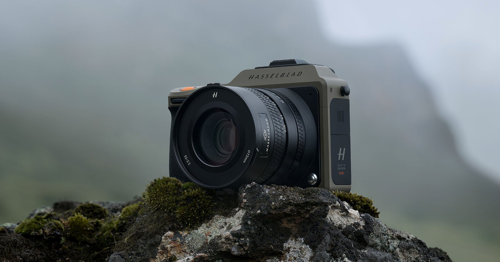 A Hasselblad camera is positioned on a moss-covered rock outdoors against a foggy, mountainous backdrop. The camera's design is sleek and modern, featuring a large lens and branding that reads Hasselblad. The setting gives a rugged and natural feel.