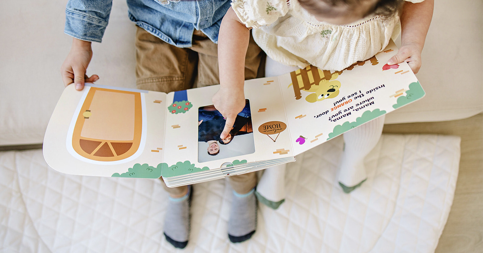 Two young children sit side by side on a quilt, looking at an open illustrated book. The page features colorful shapes and drawings. One child is pointing to a picture on the page. The children are wearing casual clothing and socks.