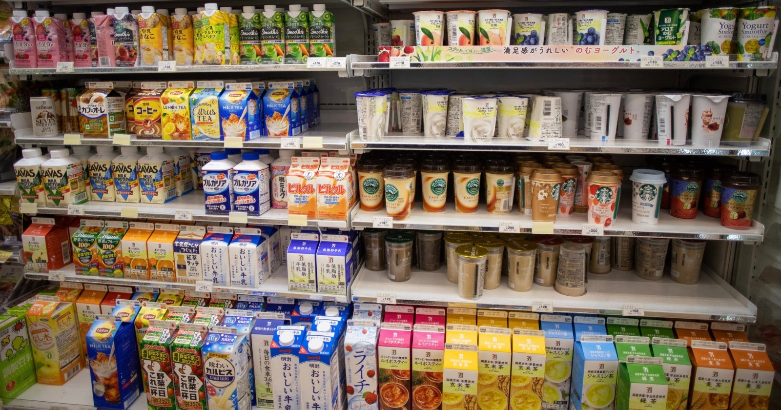 Shelves in a Japanese convenience store display a variety of colorful beverages, including juices, milks, teas, and coffees in cartons, bottles, and cans, neatly organized by type and brand.