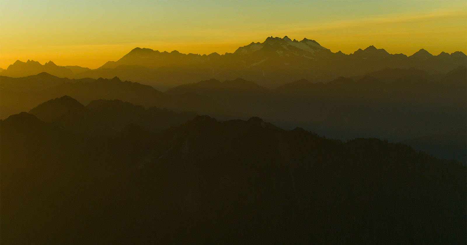 A serene mountain range at sunset, with layers of dark hills in the foreground fading into the distance. The sky is gradient tones of yellow and orange, with the highest peaks silhouetted against the horizon, creating a tranquil and picturesque landscape.