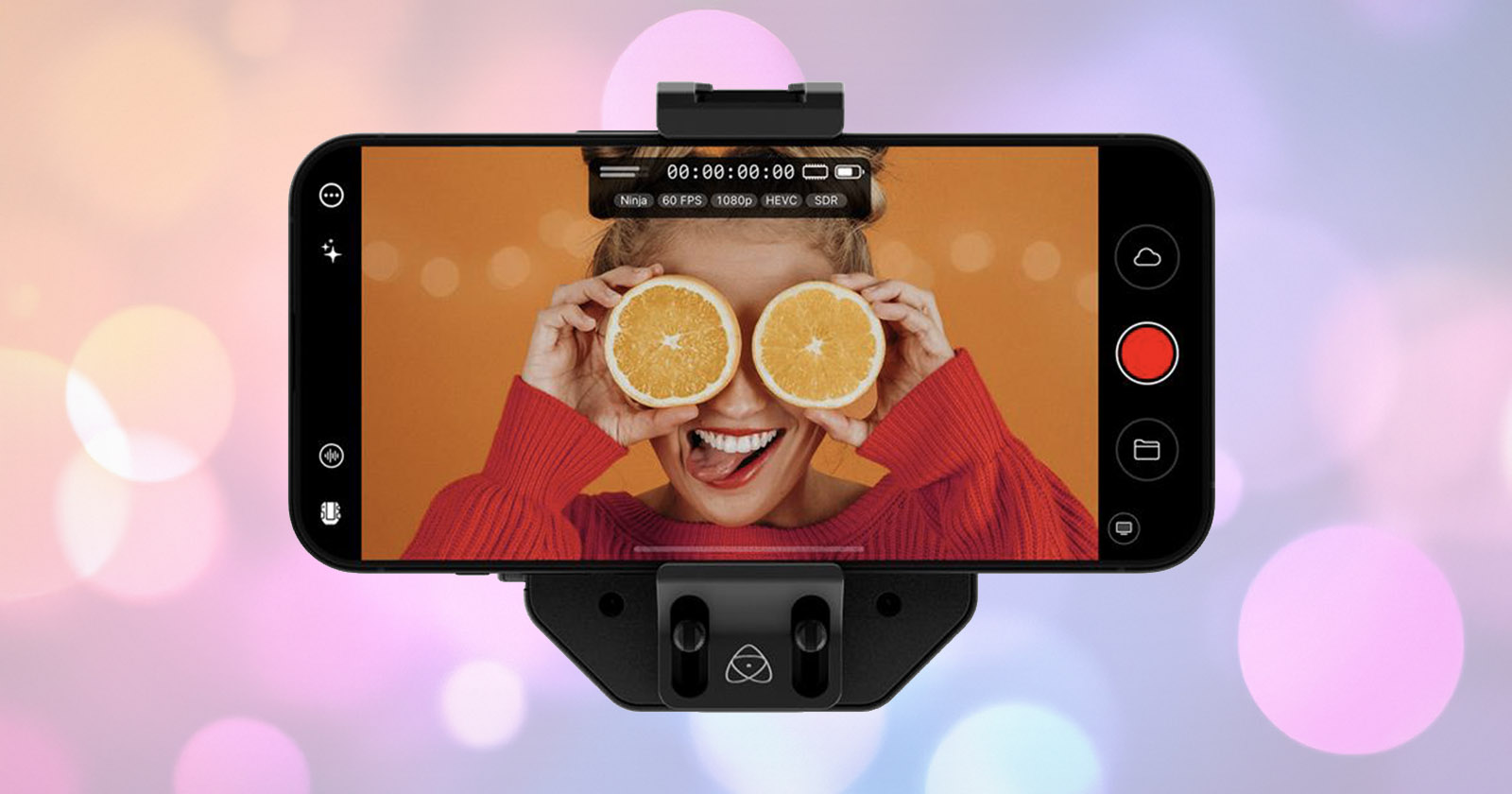 A smartphone on a mount displays a woman holding two orange halves over her eyes and smiling, with an orange background, while the phone screen shows camera controls.