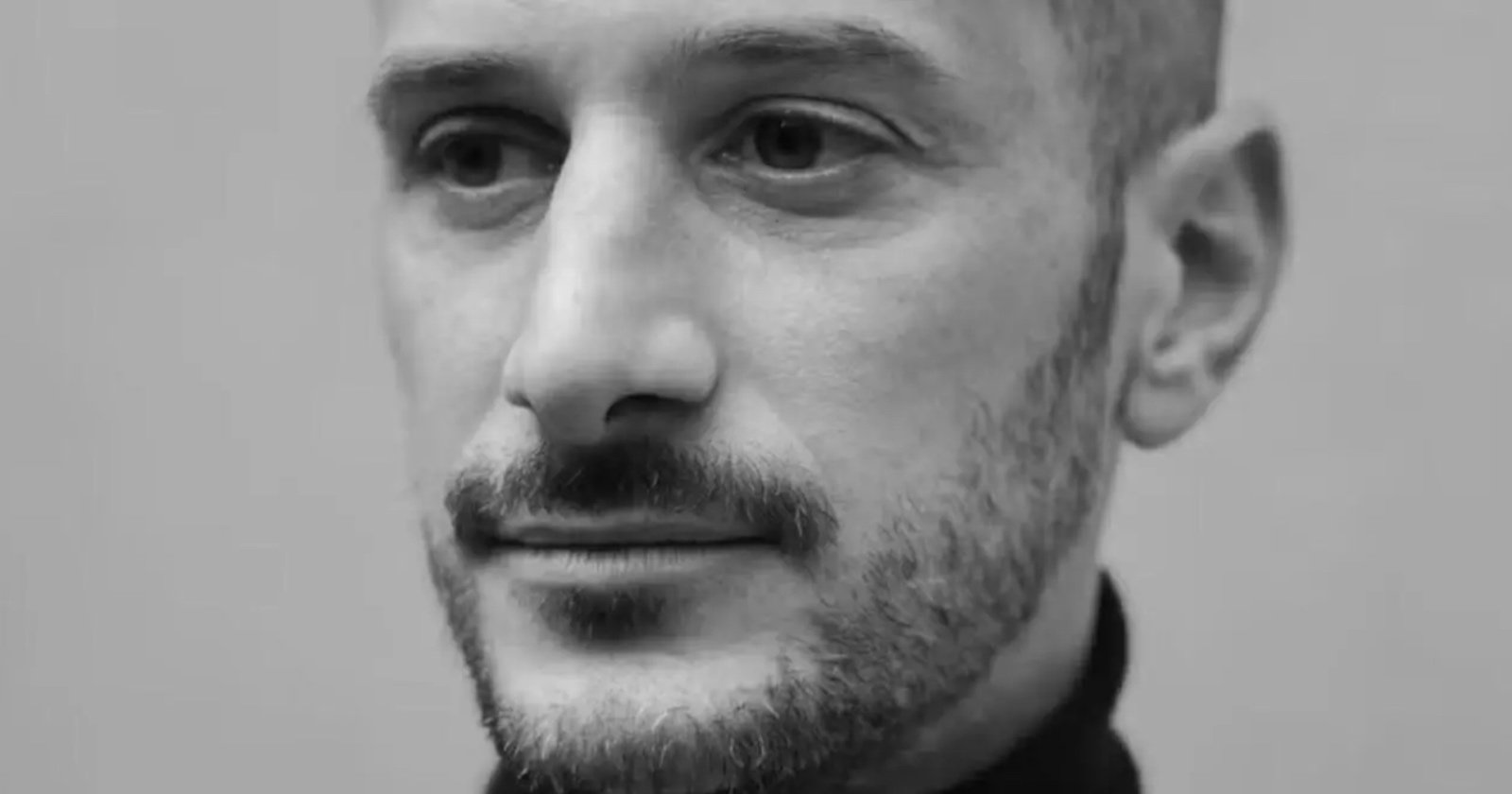 A black-and-white close-up photo of a man with a trimmed beard and mustache, wearing a dark shirt, looking slightly to the side with a neutral expression.