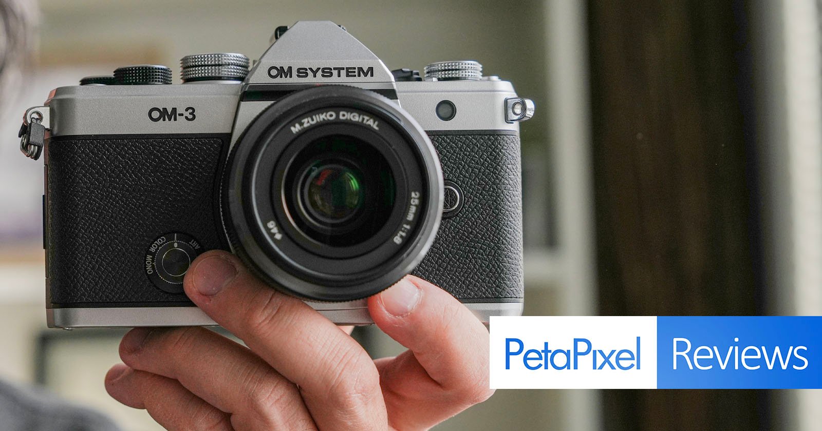 Person holding a vintage silver and black OM-3 camera with a M.Zuiko Digital lens. The image features a PetaPixel Reviews graphic in the bottom right corner. Background is blurred.