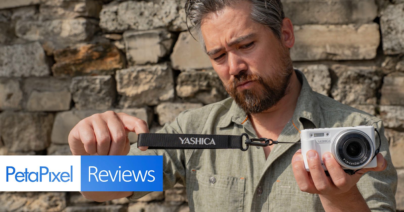 A man in a green shirt holds a white Yashica camera and camera strap outdoors, examining the strap closely. The background is a stone wall. PetaPixel Reviews is overlaid at the bottom left.