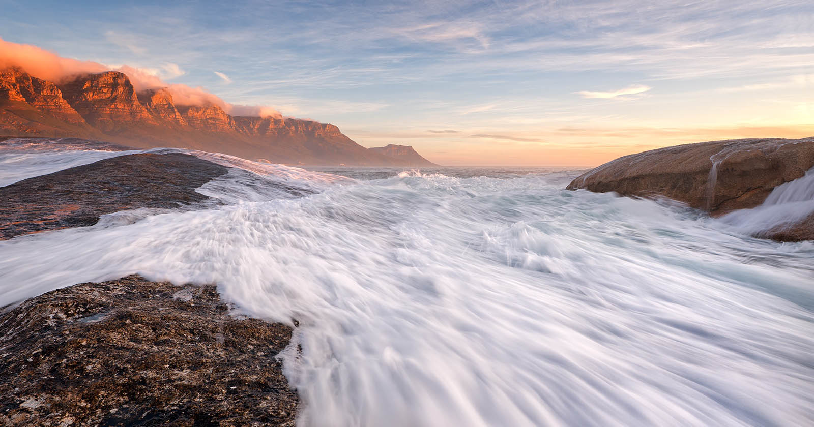 A dynamic seascape showing white waves crashing against dark rocky shorelines. In the background, mountains are bathed in the warm glow of a setting sun under a partly cloudy sky. The scene evokes a sense of power and serenity.