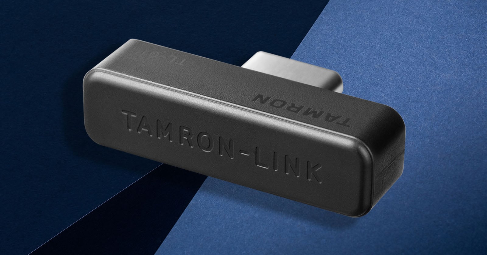 A black rectangular device labeled TAMRON-LINK and TAMRON rests on a dark and light blue geometric background.