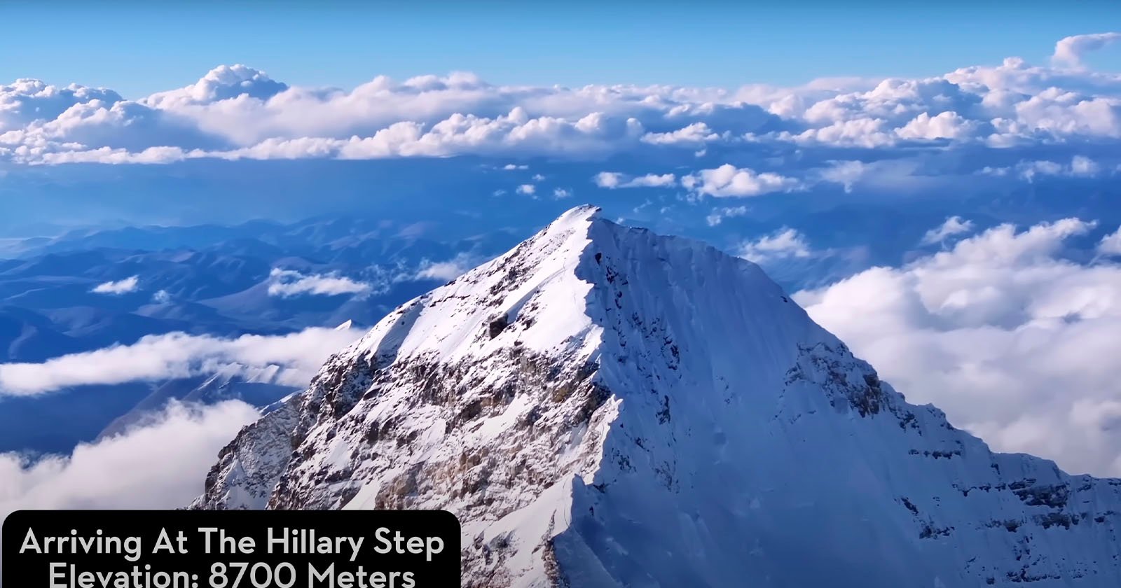 A snow-covered mountain peak rises sharply against a blue sky filled with scattered clouds. The caption reads, Arriving at The Hillary Step - Elevation: 8700 Meters. The mountain is part of a rugged, vast landscape with distant mountain ranges visible beyond.