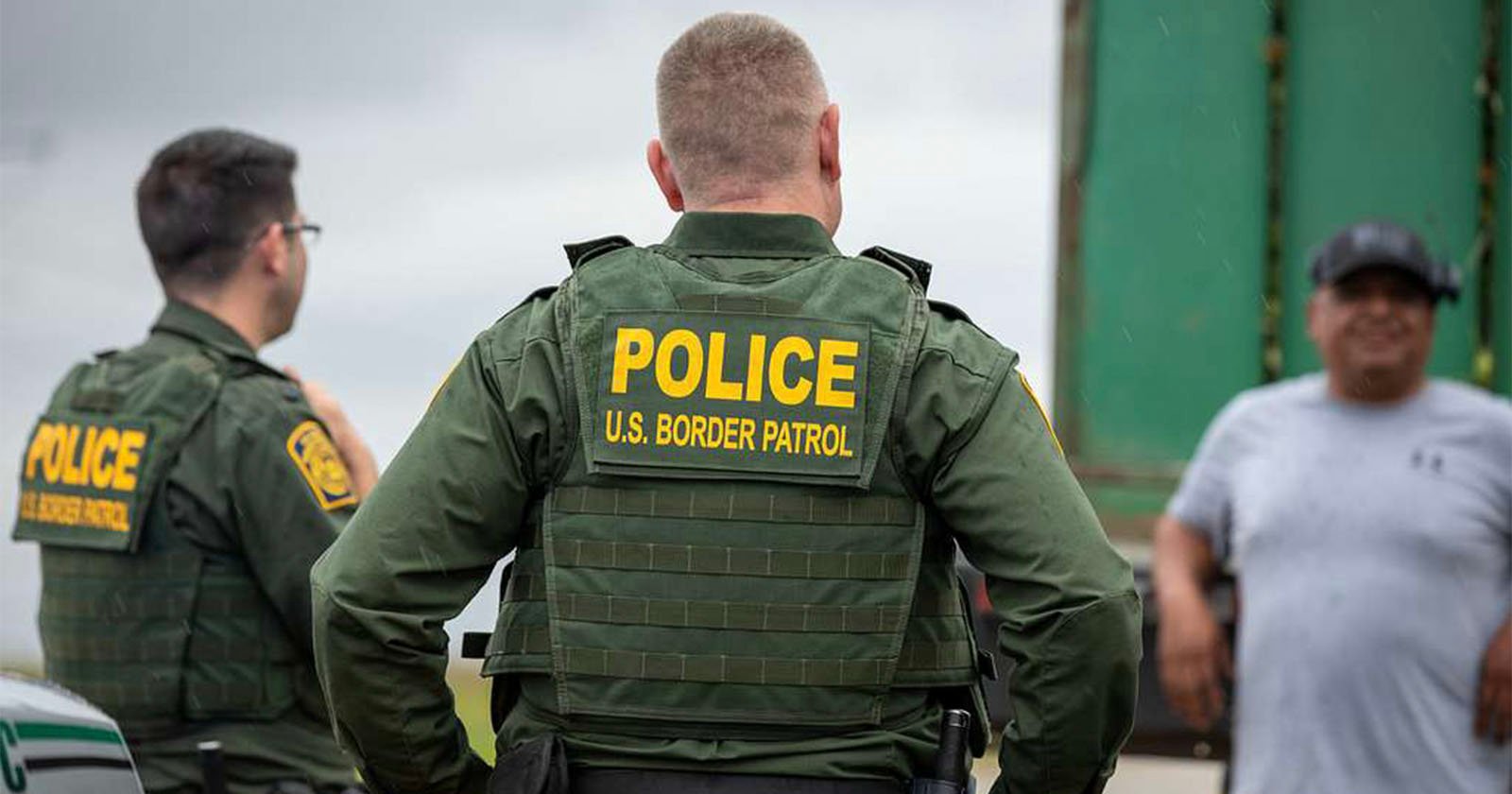Two U.S. Border Patrol officers in green uniforms face away, wearing vests with POLICE U.S. BORDER PATROL in yellow. They stand near a man in a gray shirt and cap, with a green truck visible in the background.
