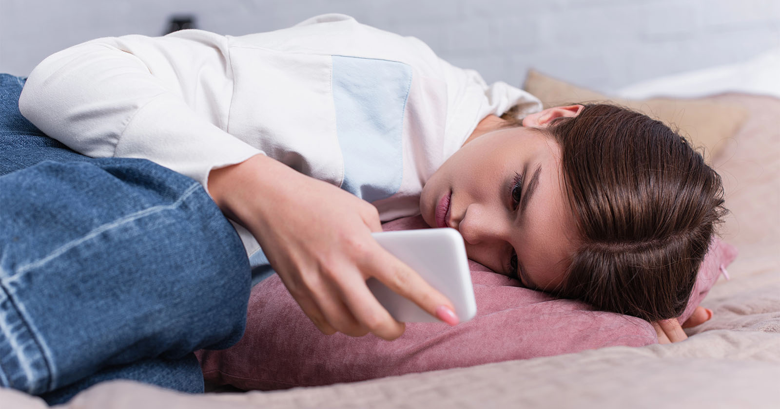 A young woman lies on her side on a bed, resting her head on a pillow while looking at a smartphone. She appears to be thoughtful or sad, wearing a white shirt and jeans.