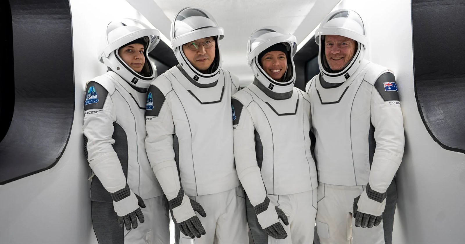 Four astronauts in white and gray SpaceX suits stand inside a spacecraft, smiling and facing the camera. They wear helmets with clear visors and are positioned closely together in a narrow passageway.