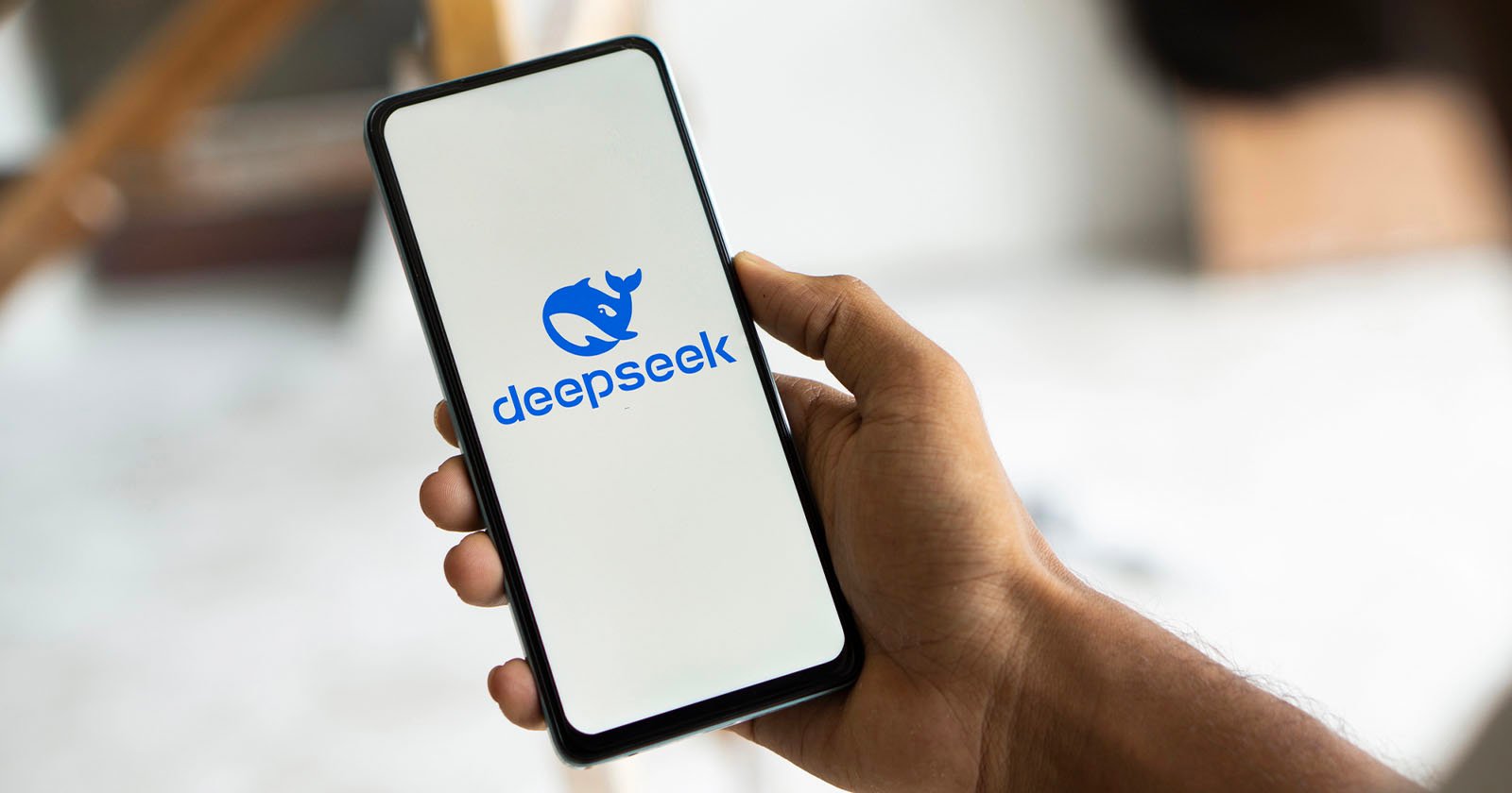 A person holds a smartphone displaying the deepseek logo, featuring a stylized rhinoceros head. The screen background is white, and the setting appears to be indoors with a blurred background.