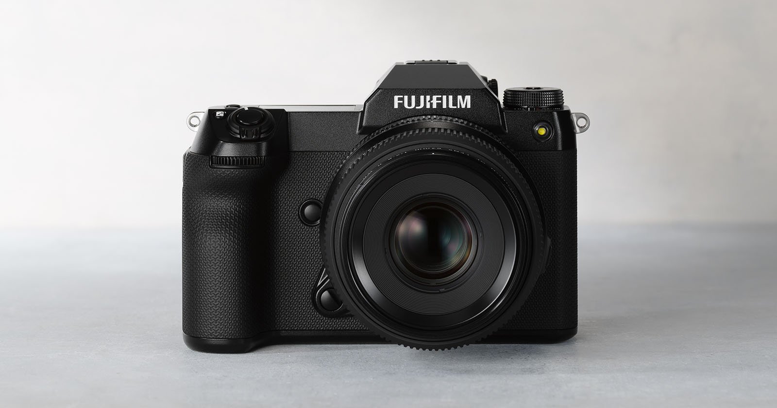 Front view of a black Fujifilm professional digital camera placed on a light gray surface. The camera features a prominent lens and detailed buttons on the left side of its body. The brand name Fujifilm is clearly visible on the top of the camera.