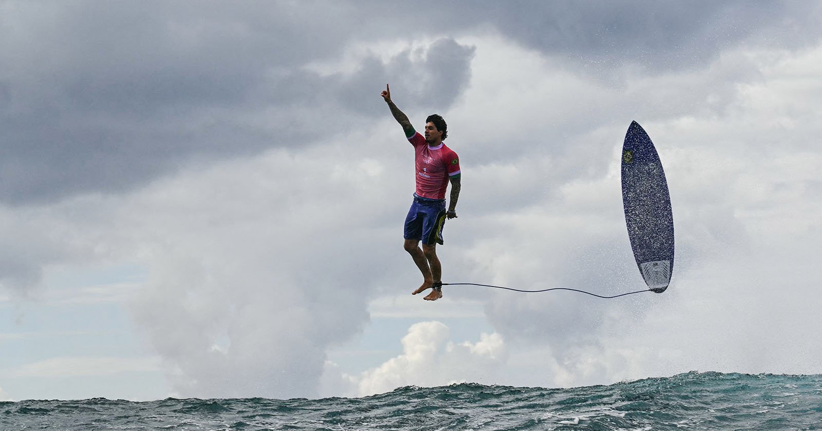 A surfer, clad in a pink shirt and blue shorts, leaps high above the ocean waves, the surfboard tethered to him by a leg rope. He points skyward with one hand under a cloudy sky.