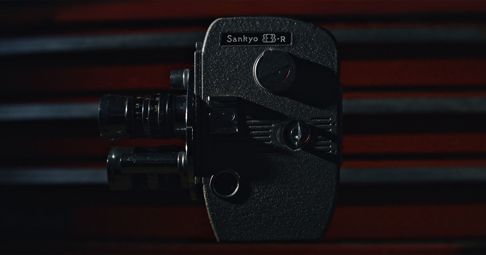 A vintage Sankyo 8-R movie camera is shown close-up against a dark background with red horizontal lines, highlighting its textured body and lens details.