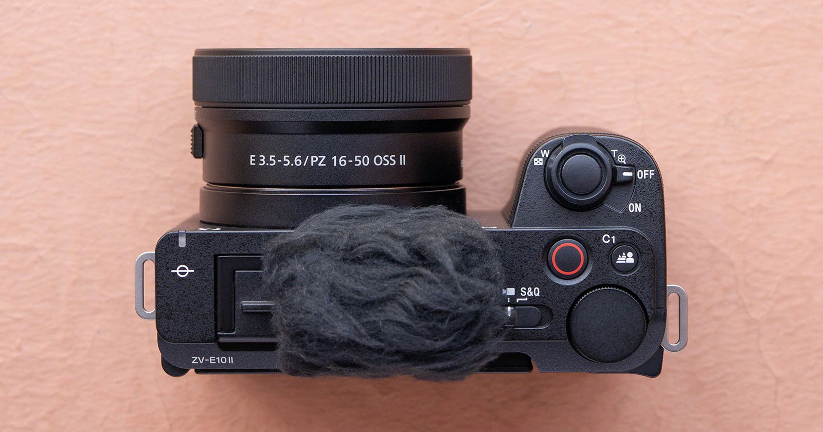 A black camera with a rubberized grip and a lens labeled E 3.5-5.6/PZ 16-50 OSS II is placed on a pink surface. The top of the camera has several buttons and dials, including a red record button, a function button labeled C1, and a power switch.