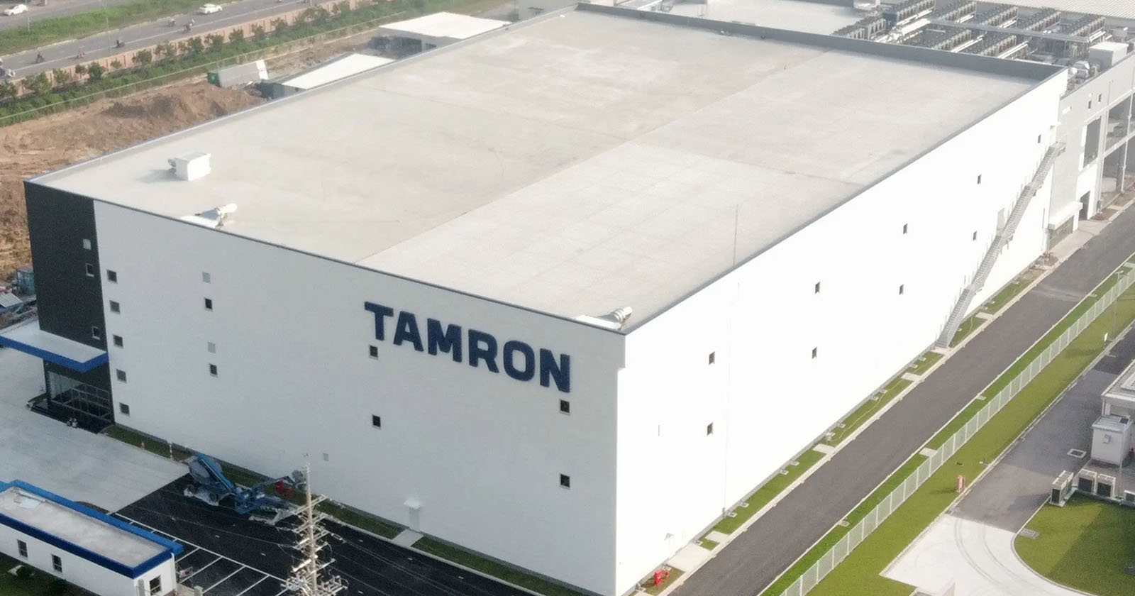 Aerial view of a large, white industrial building with TAMRON written in bold blue letters on the side. The building is surrounded by roads and greenery, featuring a paved area with vehicles nearby.
