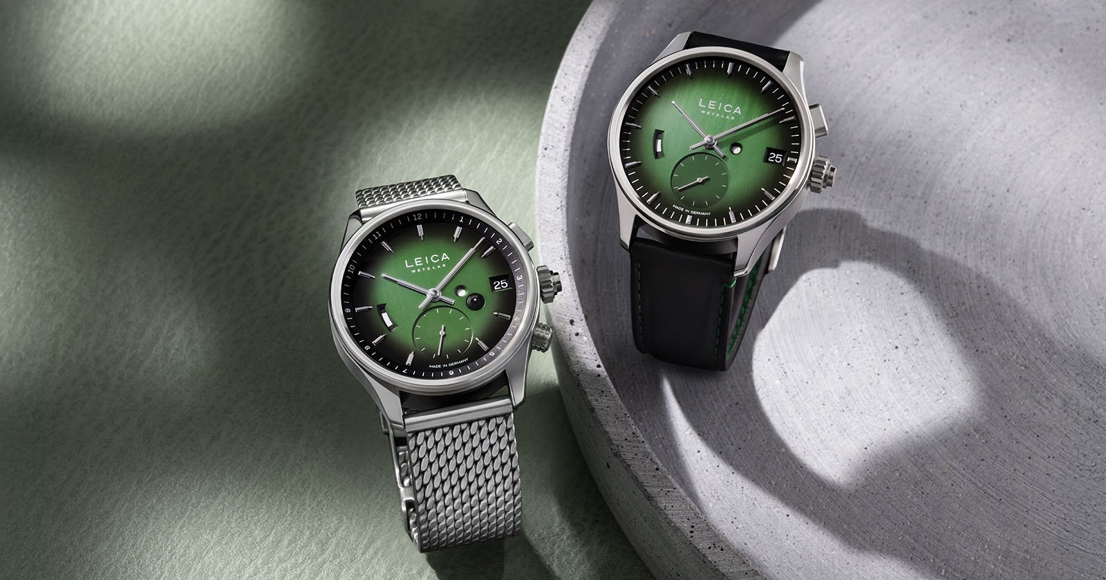 Two luxury Leica watches with green dials; one has a metal mesh band and the other a black leather strap. Both are placed on a green and gray textured surface with soft lighting and shadows.