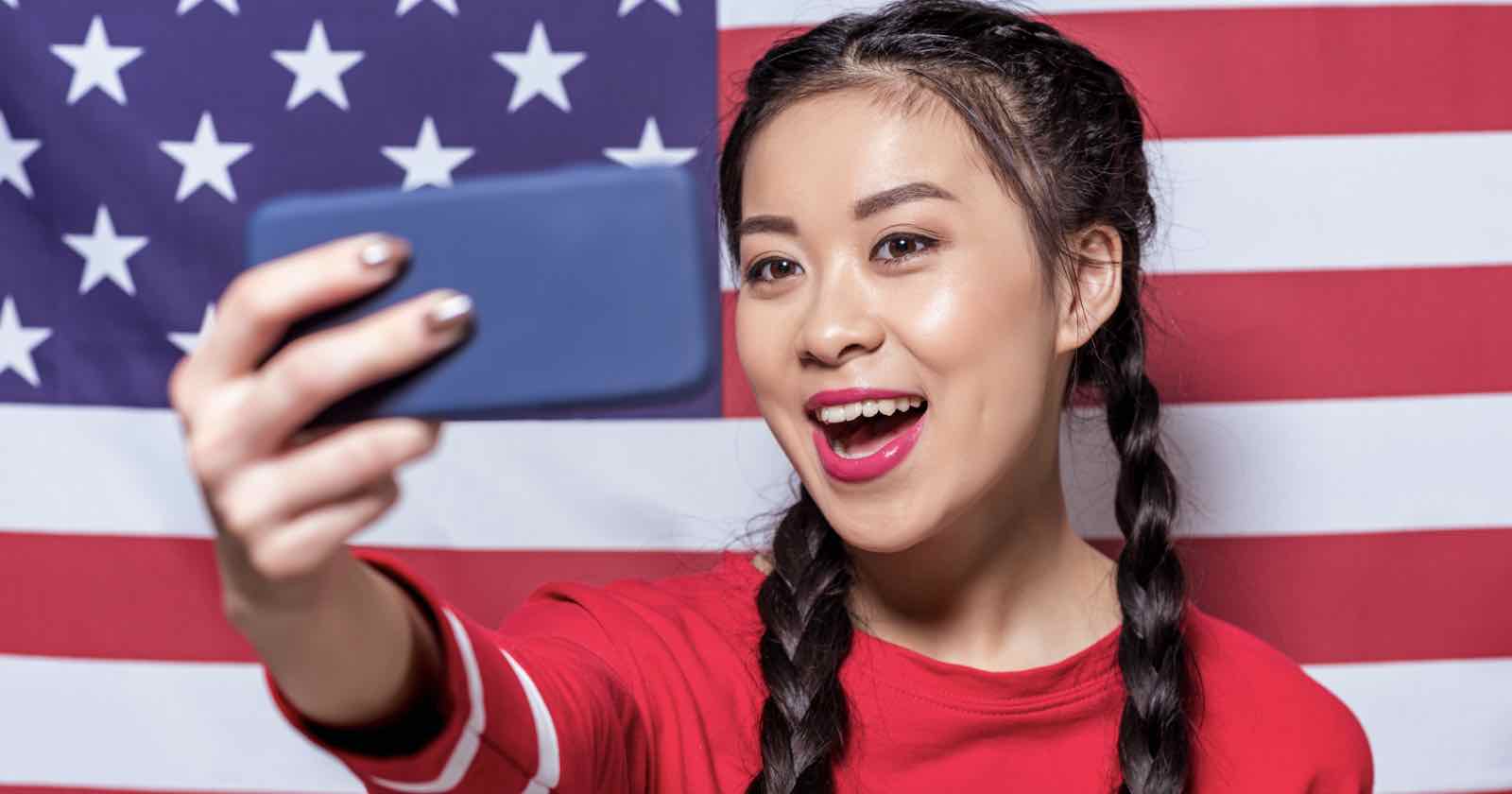 A woman with braided hair smiles while taking a selfie with a blue smartphone. She is wearing a red top and standing in front of an American flag.
