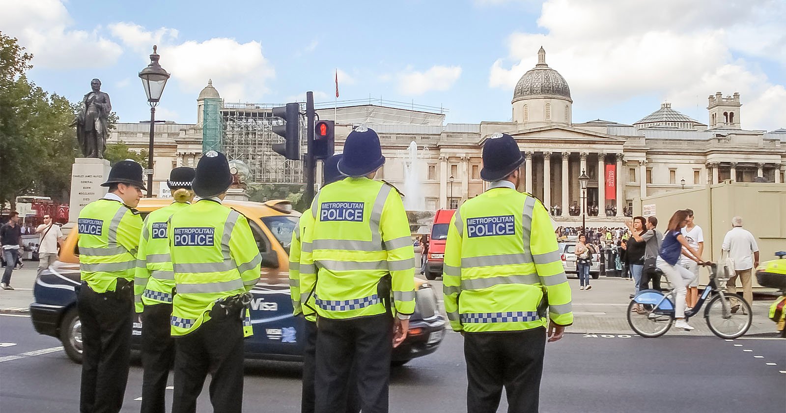 Four Metropolitan Police officers in high-visibility jackets stand on a street corner facing away, observing a busy area. In the background, there is a historic building with columns and a dome, and several pedestrians, cars, and cyclists are visible.