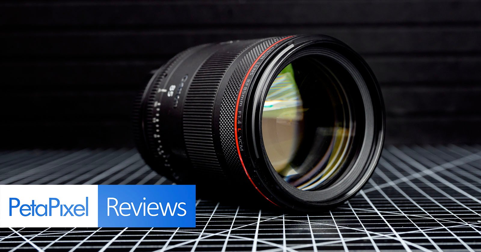 A camera lens rests on a black grid-patterned surface with a dark, lined background. The words PetaPixel Reviews appear in blue and white boxes at the bottom left corner.