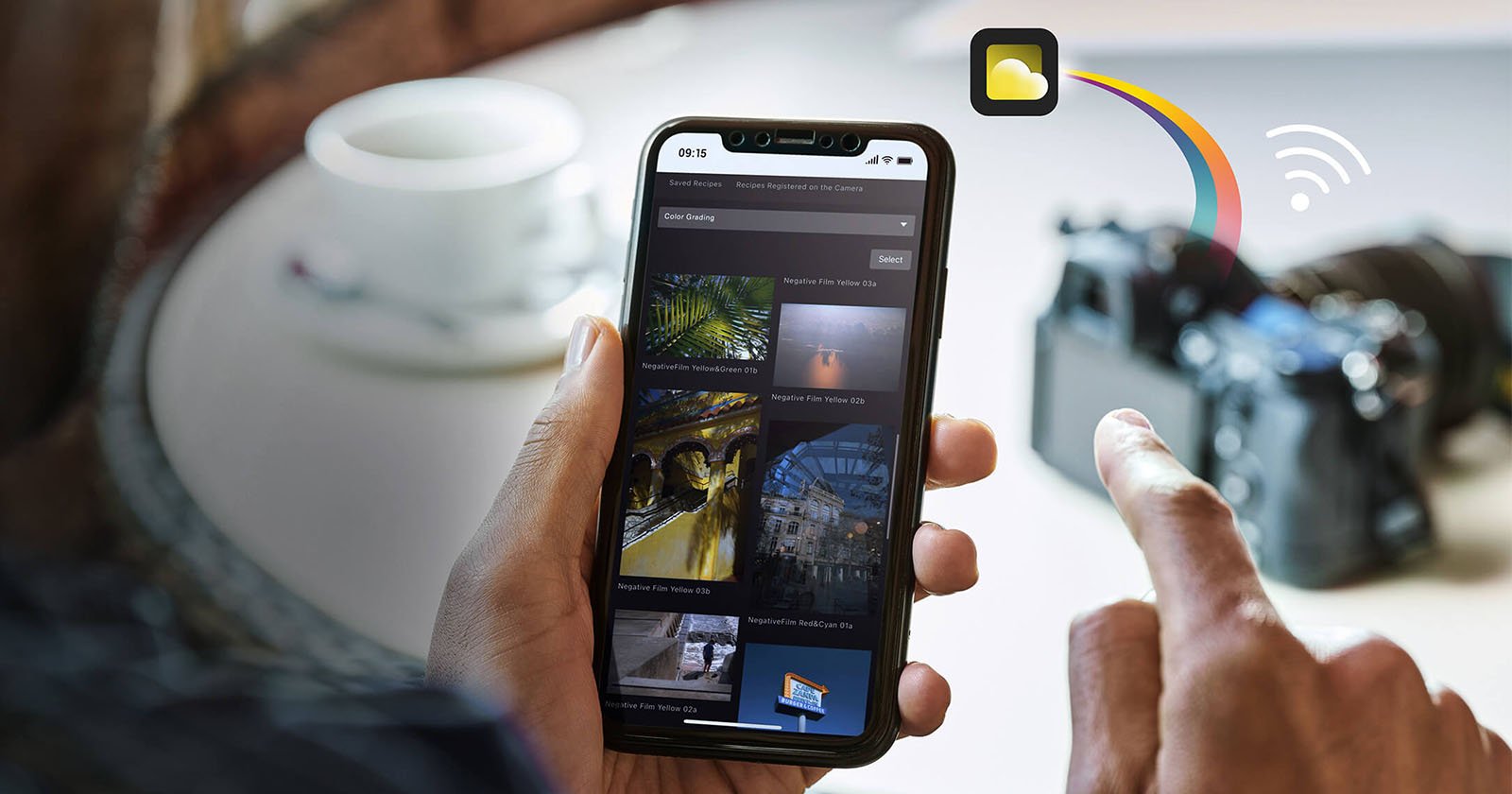 A person holds a smartphone showing a gallery of images with options for selection. Next to the phone is a camera on a table. An icon of a yellow cloud with blue lines indicates wireless image transfer from the camera to the phone. A coffee cup is in the background.