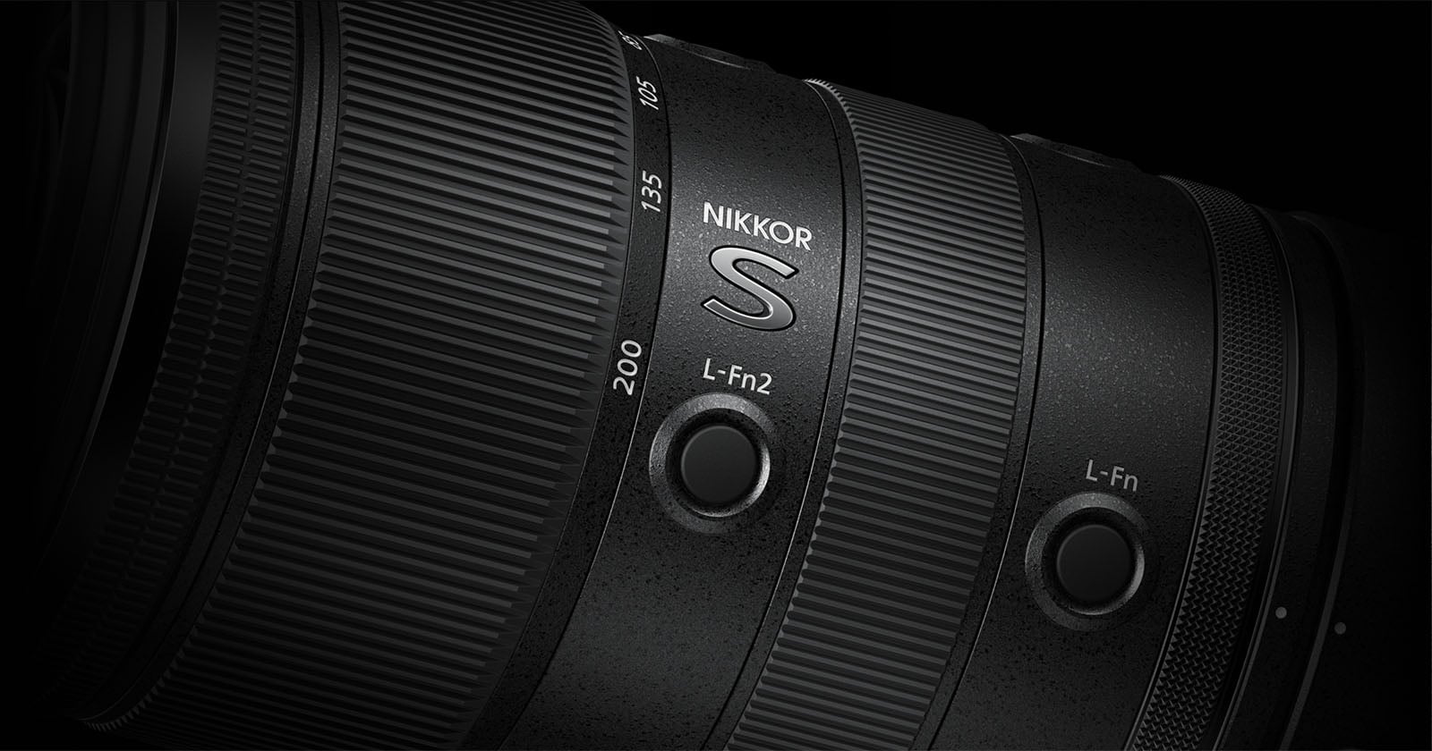 Close-up of a black Nikkor S camera lens, showing detailed texture, focal length markings (135, 200), and two customizable L-Fn buttons on the lens barrel against a dark background.