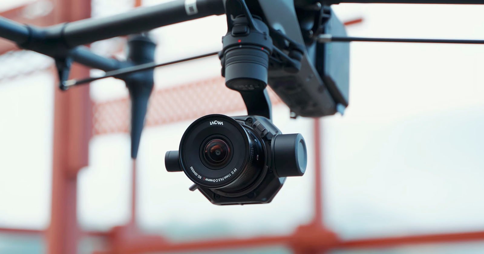 A close-up of a black camera mounted on a drone, with a blurred bridge structure in the background. the camera lens has a visible brand name.